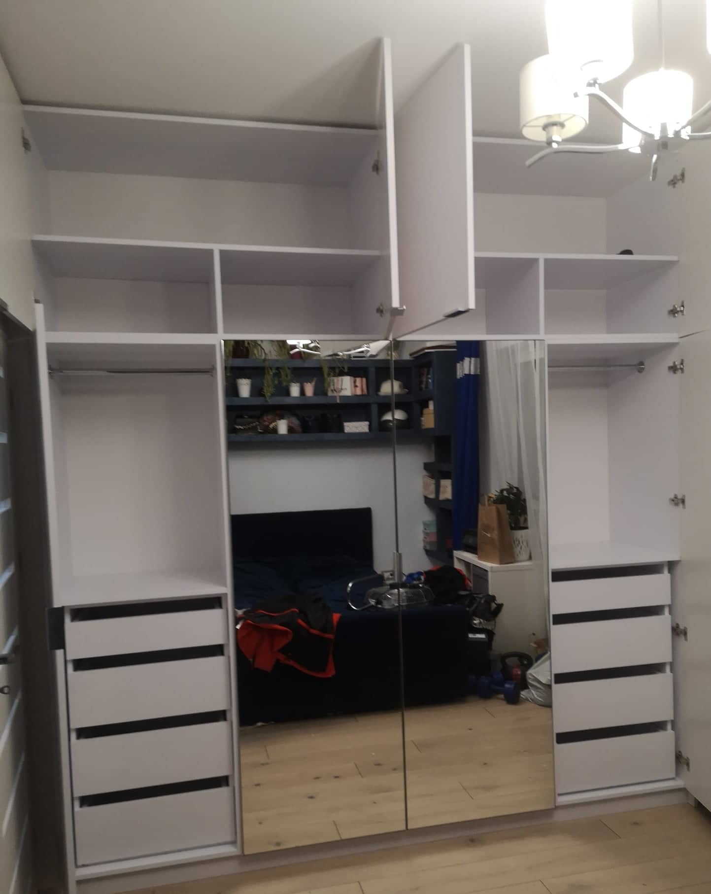 Biała szafa wnękowa z lustrzanymi drzwiami odbijającymi fragment pokoju z niebieską sofą i półkami na ścianie.