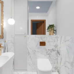 Nowoczesna łazienka z marmurowymi ścianami i podłogą, wisząca toaleta, umywalka nablatowa z baterią w kolorze złotym, lustro w ozdobnej ramie i szafka w zabudowie.