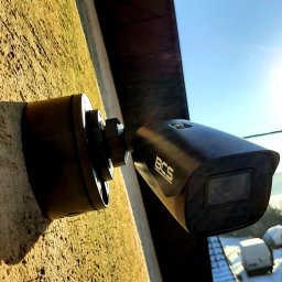 Electro Zych - Czarna kamera monitoringu BCS zamontowana na szorstkiej, oświetlonej słońcem, żółtej ścianie budynku. Widoczne przewody elektryczne na tle błękitnego nieba.