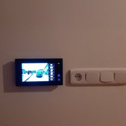 Electro Zych - Zamontowany wideodomofon z widokiem na podjazd, obok gniazdko elektryczne i włączniki światła na kremowej ścianie.