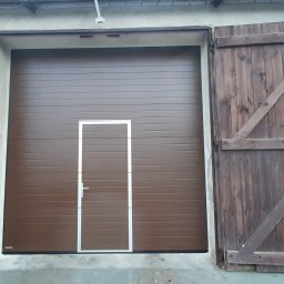 Electro Zych - Brązowa segmentowa brama garażowa z drzwiami serwisowymi pośrodku, otoczona starymi, drewnianymi wrotami z widocznymi zawiasami i zasuwami, osadzona w szarej, lekko uszkodzonej elewacji budynku.