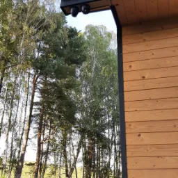 Dwie czarne kamery monitoringu zamontowane pod okapem drewnianego domu z widokiem na ogród i drzewa.