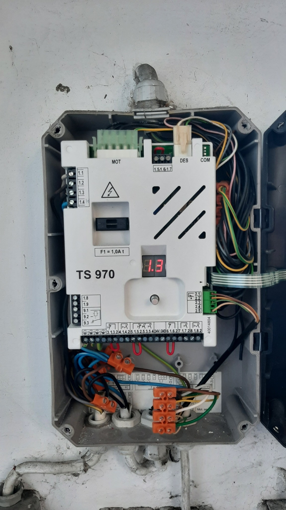 Otwarta skrzynka sterownicza TS 970 z widocznymi przewodami, złączkami i elementami elektronicznymi, zamontowana na szarej ścianie.