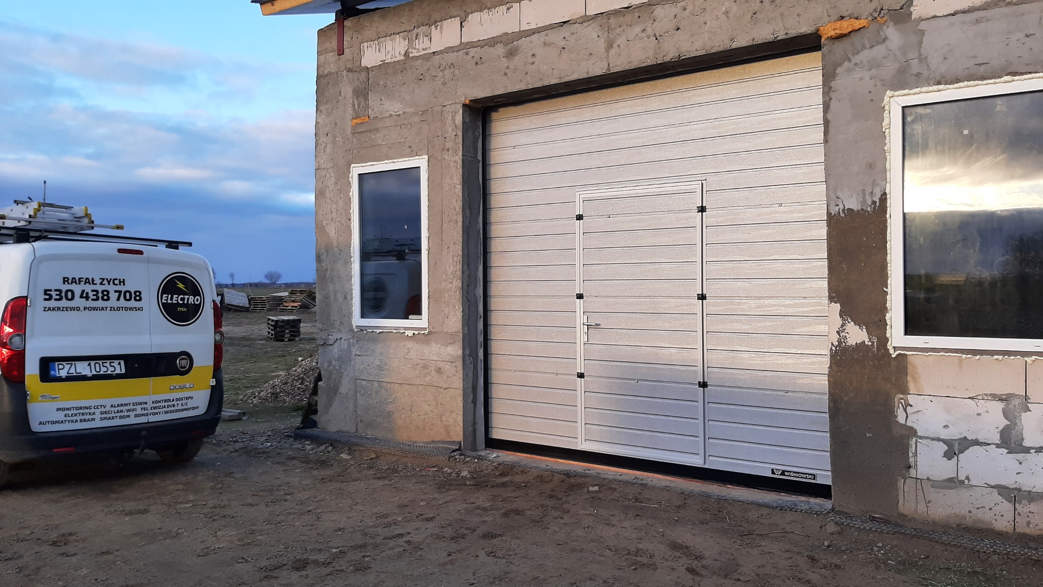 Szara brama garażowa segmentowa z drzwiami wejściowymi, zamontowana w surowym budynku, obok zaparkowany biały samochód dostawczy z logo firmy i numerem telefonu.