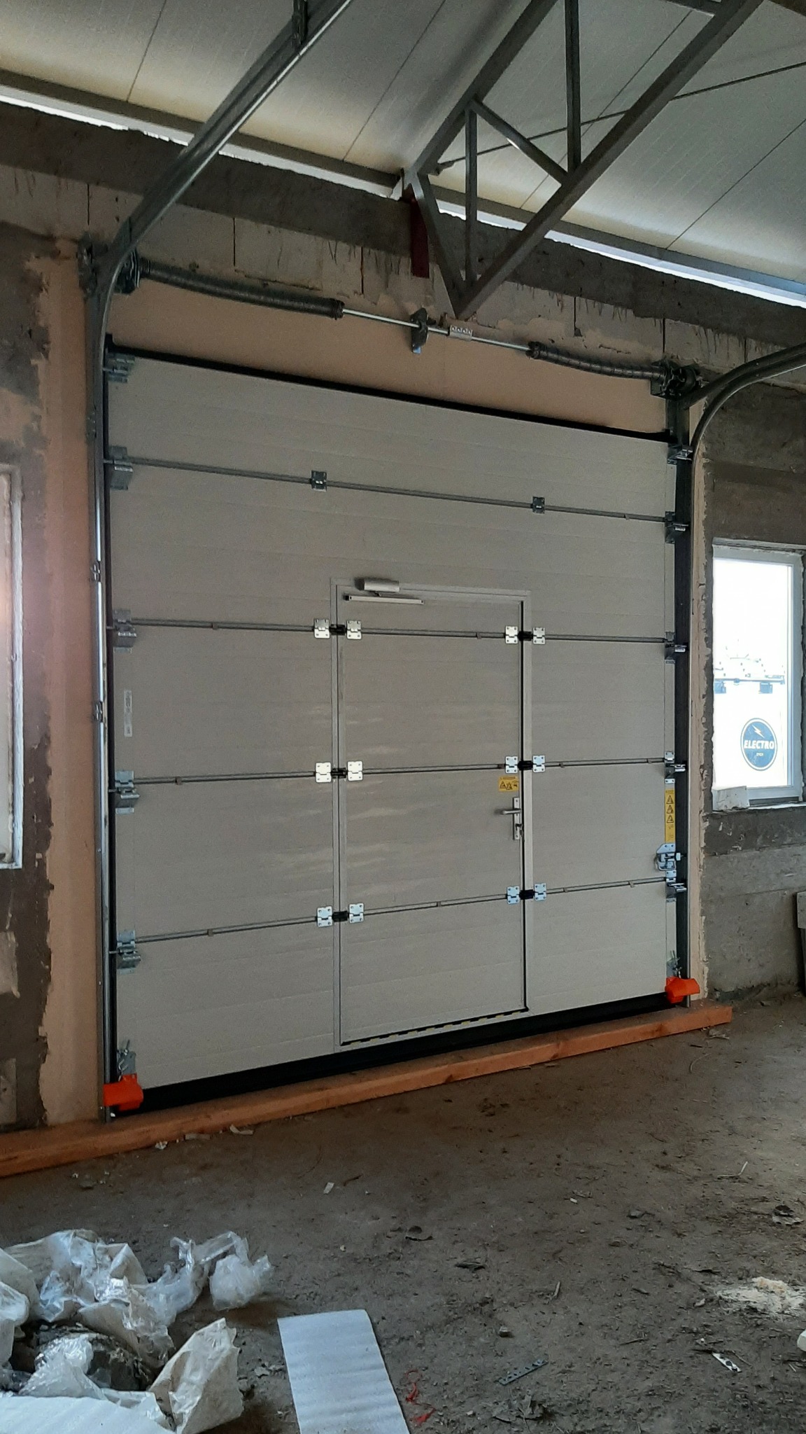 Zamknięta segmentowa brama garażowa z drzwiami przejściowymi w Zakrzewie, widoczne pomarańczowe ograniczniki i surowy stan wykończenia wnętrza.