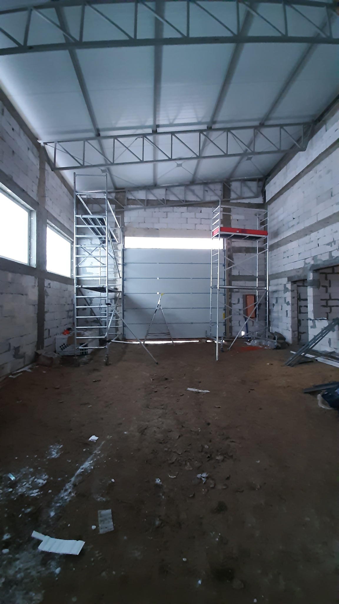 Wnętrze surowego budynku z bramą segmentową w trakcie montażu, widoczne rusztowania i surowe ściany z bloczków.