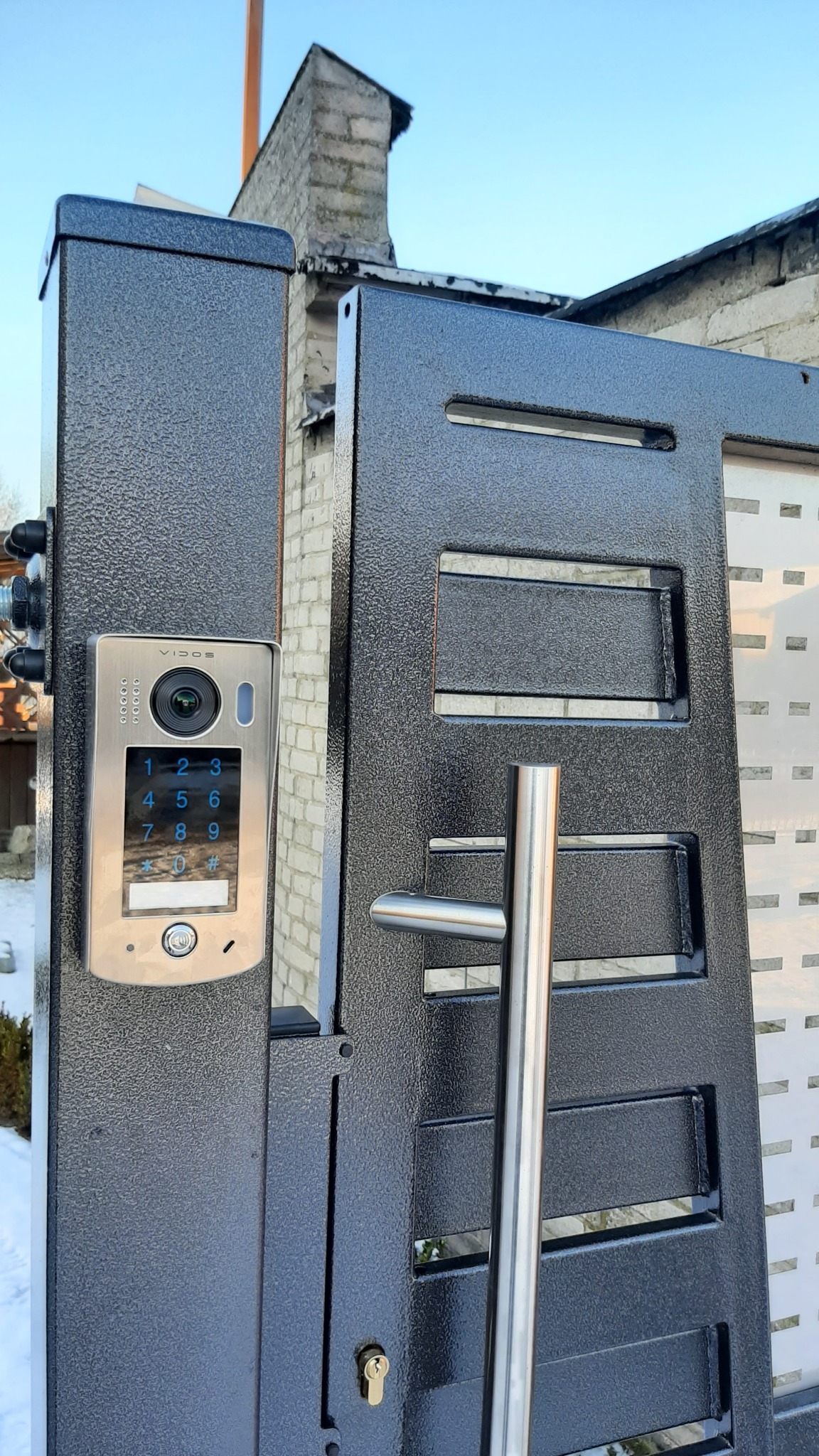 Nowoczesny wideodomofon Vidos zamontowany na metalowej bramie posesji z widocznym panelem numerycznym i kamerą.
