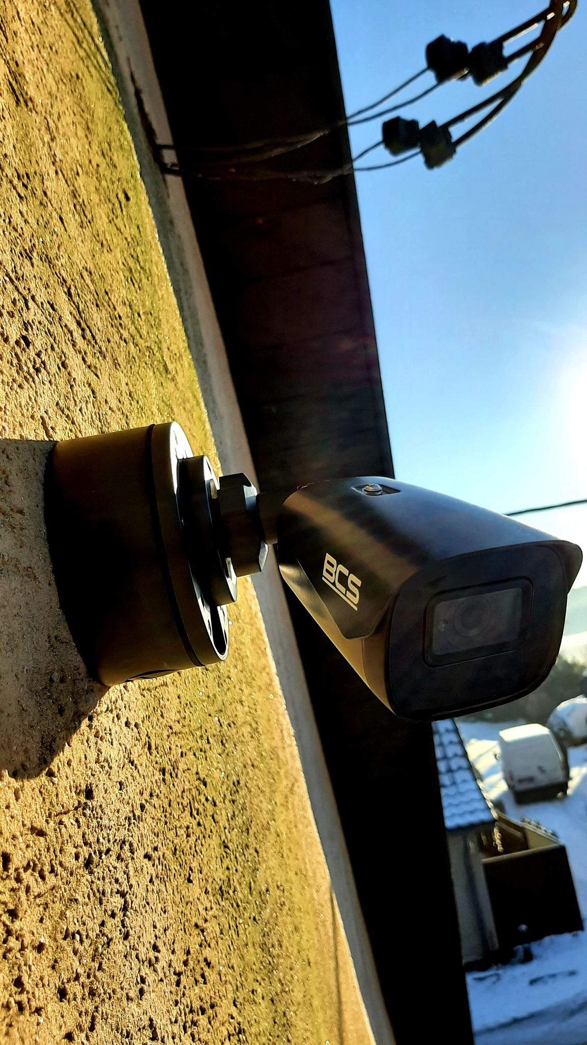 Czarna kamera monitoringu BCS zamontowana na szorstkiej, oświetlonej słońcem, żółtej ścianie budynku. Widoczne przewody elektryczne na tle błękitnego nieba.