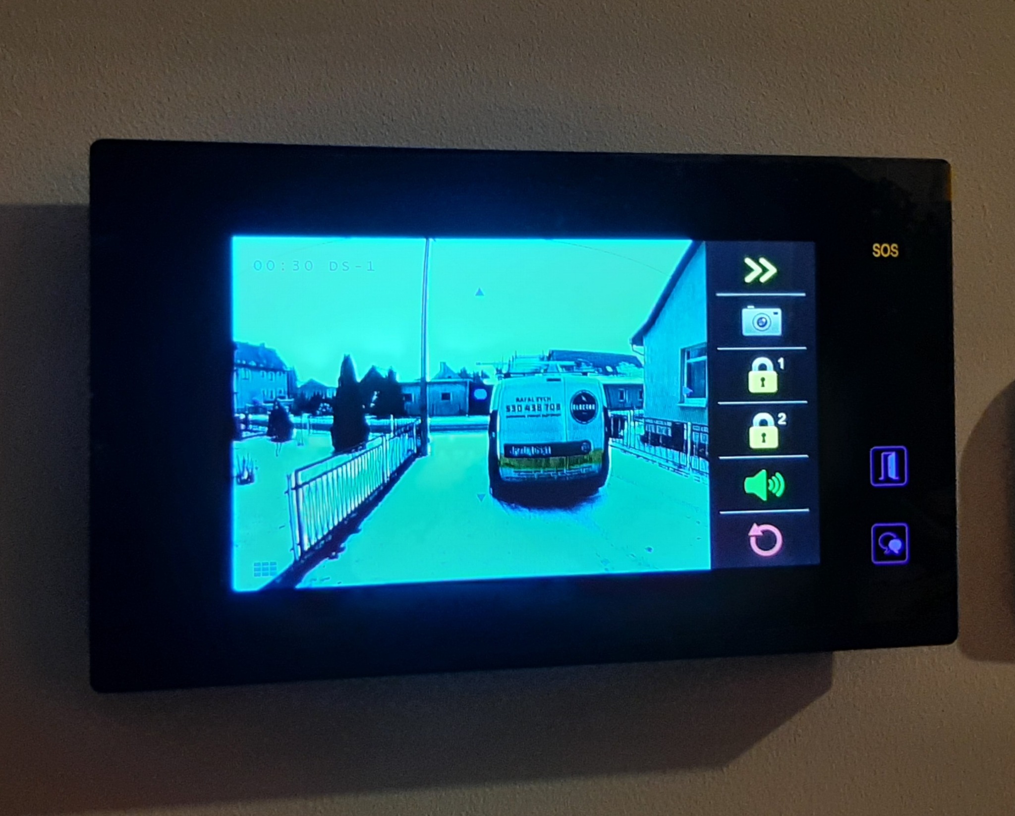 Ekran wideodomofonu wyświetlający obraz z kamery: widok na ulicę i zaparkowany samochód dostawczy z logo firmy.