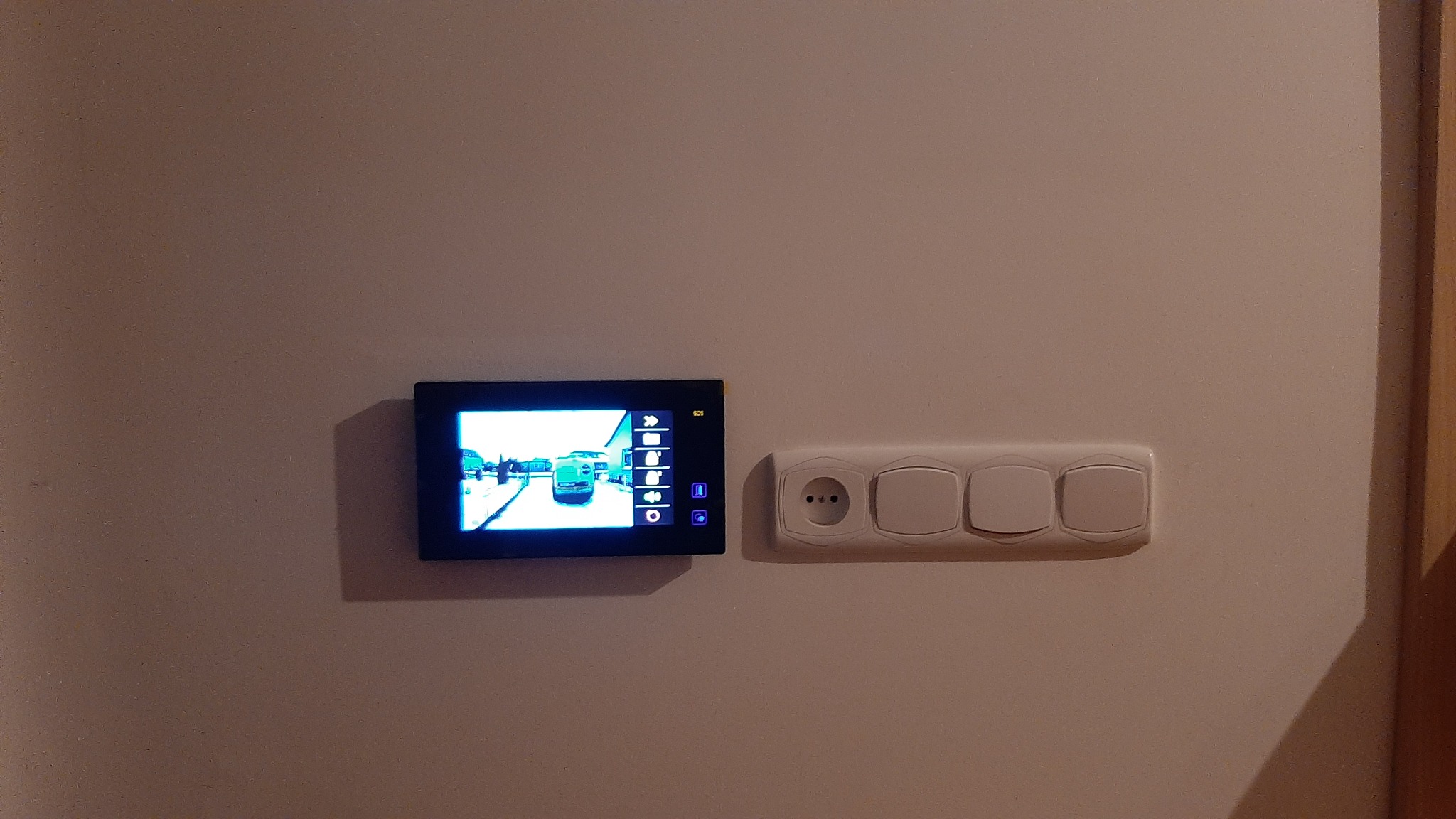 Zamontowany wideodomofon z widokiem na podjazd, obok gniazdko elektryczne i włączniki światła na kremowej ścianie.