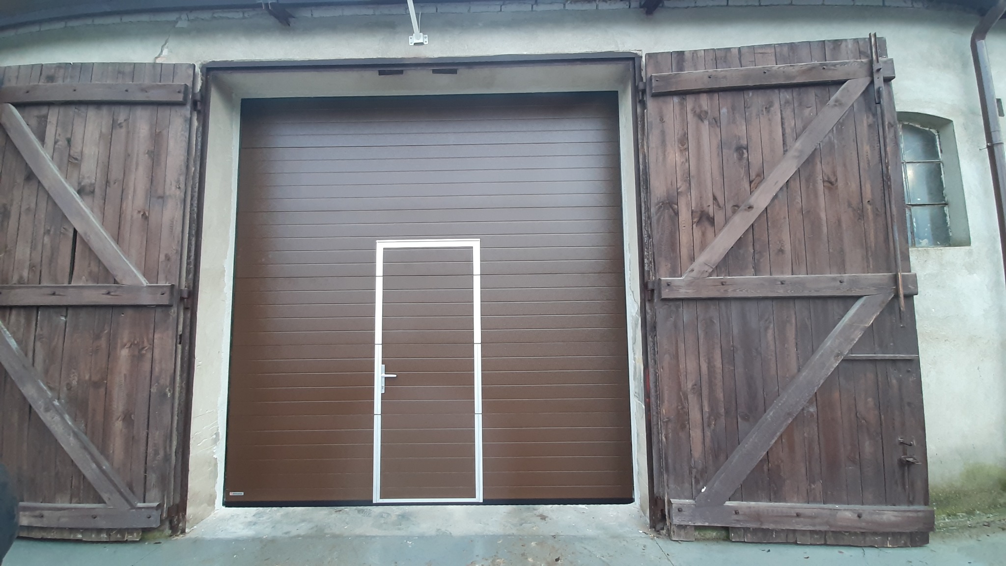 Brązowa segmentowa brama garażowa z drzwiami serwisowymi pośrodku, otoczona starymi, drewnianymi wrotami z widocznymi zawiasami i zasuwami, osadzona w szarej, lekko uszkodzonej elewacji budynku.
