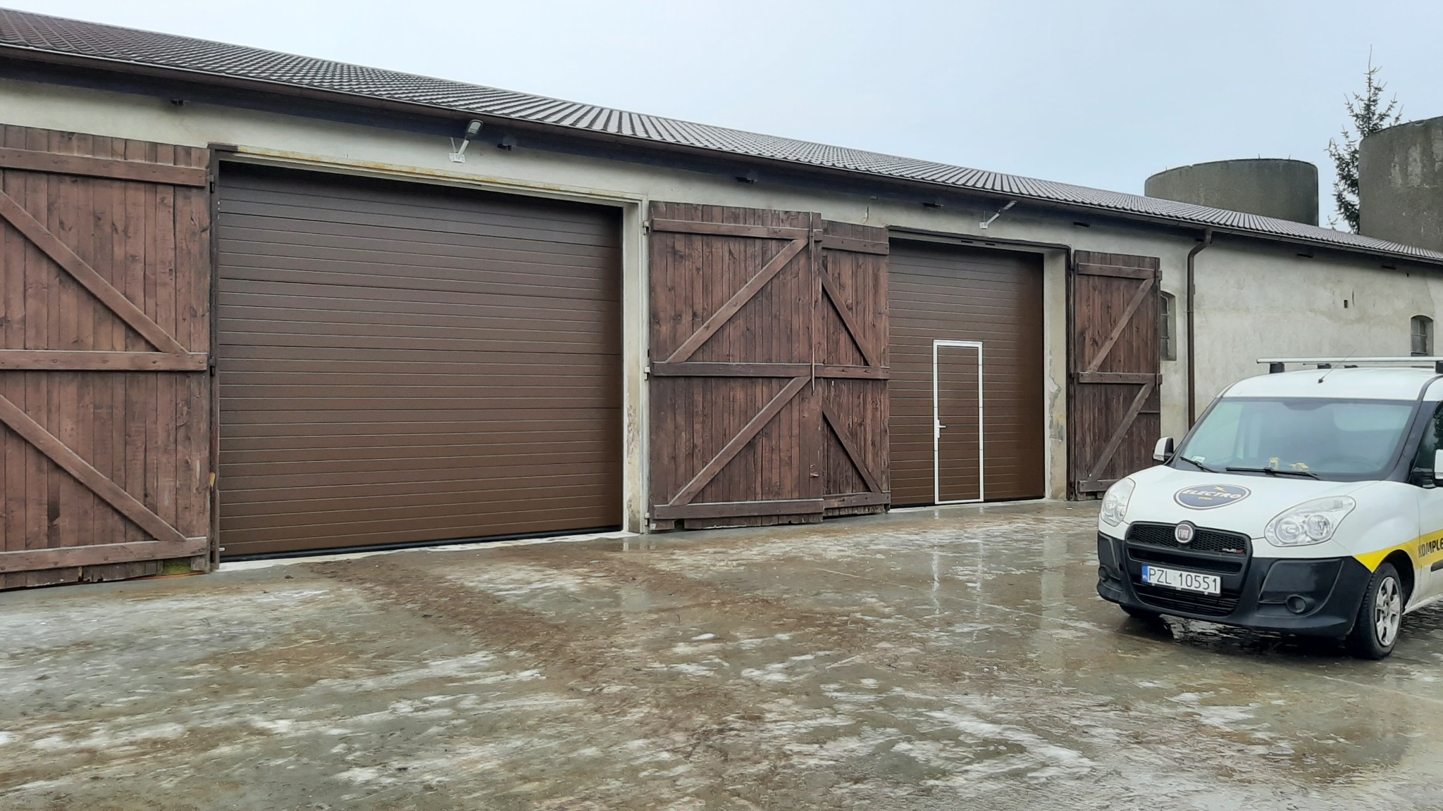 Brązowe segmentowe bramy garażowe z drewnianymi drzwiami obok, na tle budynku gospodarczego i firmowy samochód dostawczy z logo.