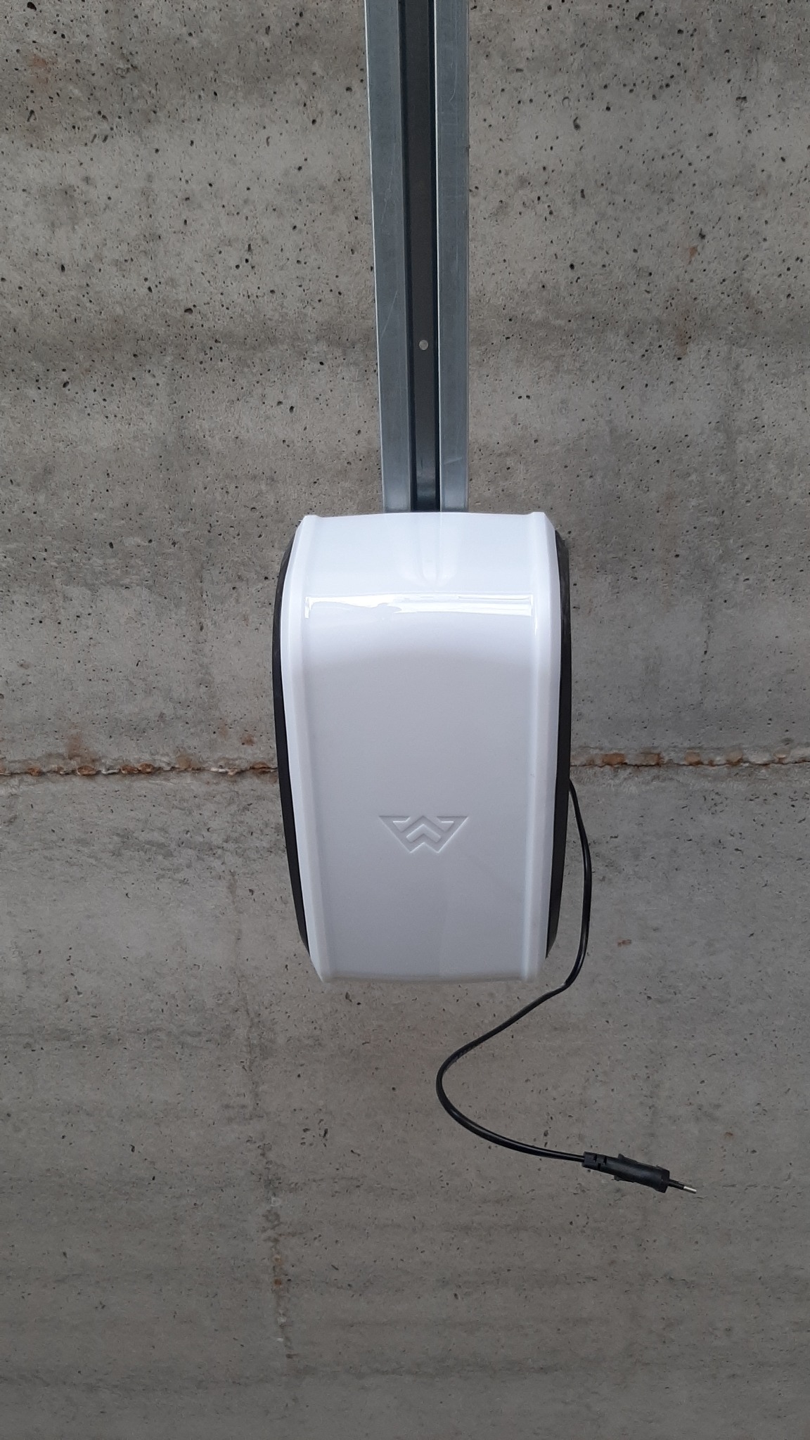 Zamontowany napęd bramy garażowej z białą obudową i widocznym logo, przymocowany do szyny montażowej pod betonowym sufitem.