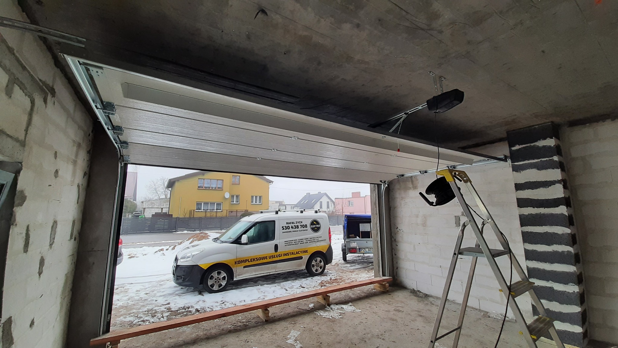 Montaż segmentowej bramy garażowej z napędem elektrycznym, widoczny samochód serwisowy firmy instalacyjnej na zewnątrz, drabina wewnątrz garażu.