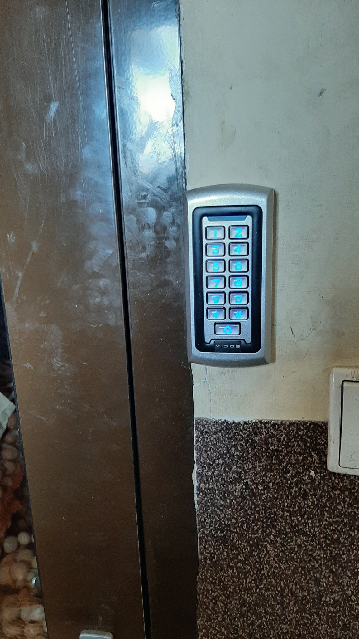 Szary panel dotykowy domofonu Vidos z podświetlanymi na niebiesko cyframi, zamontowany na pomalowanej ścianie obok metalowych drzwi.