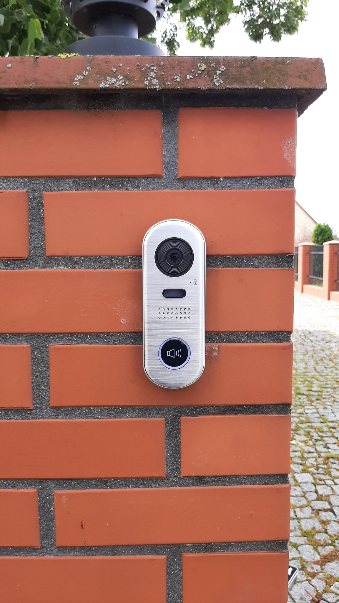 Nowoczesny, srebrny wideodomofon zamontowany na ceglanym słupku ogrodzeniowym z widoczną kamerą, głośnikiem i podświetlanym przyciskiem dzwonka.