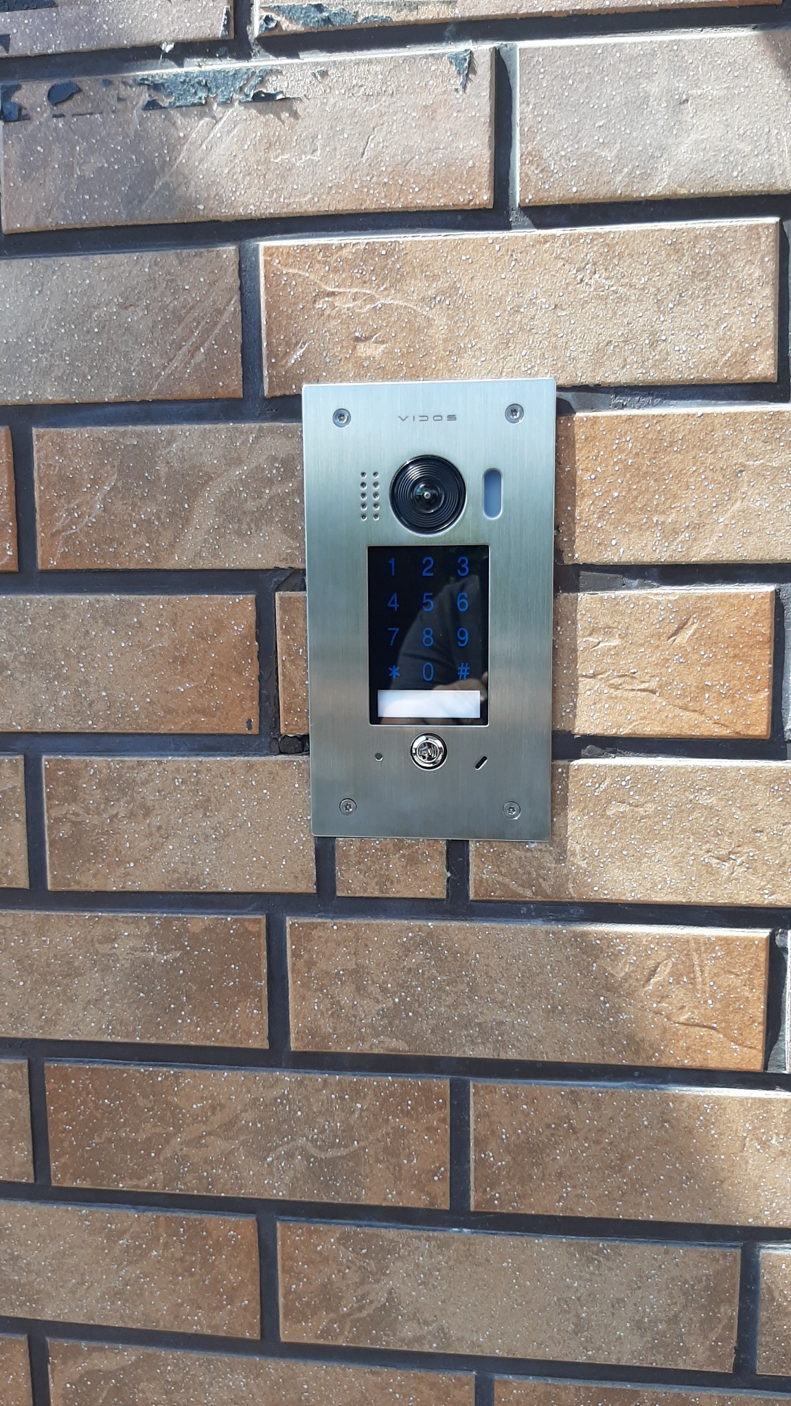 Panel wideodomofonu Vidos z klawiaturą numeryczną, zamontowany na ceglanym murze zewnętrznym.