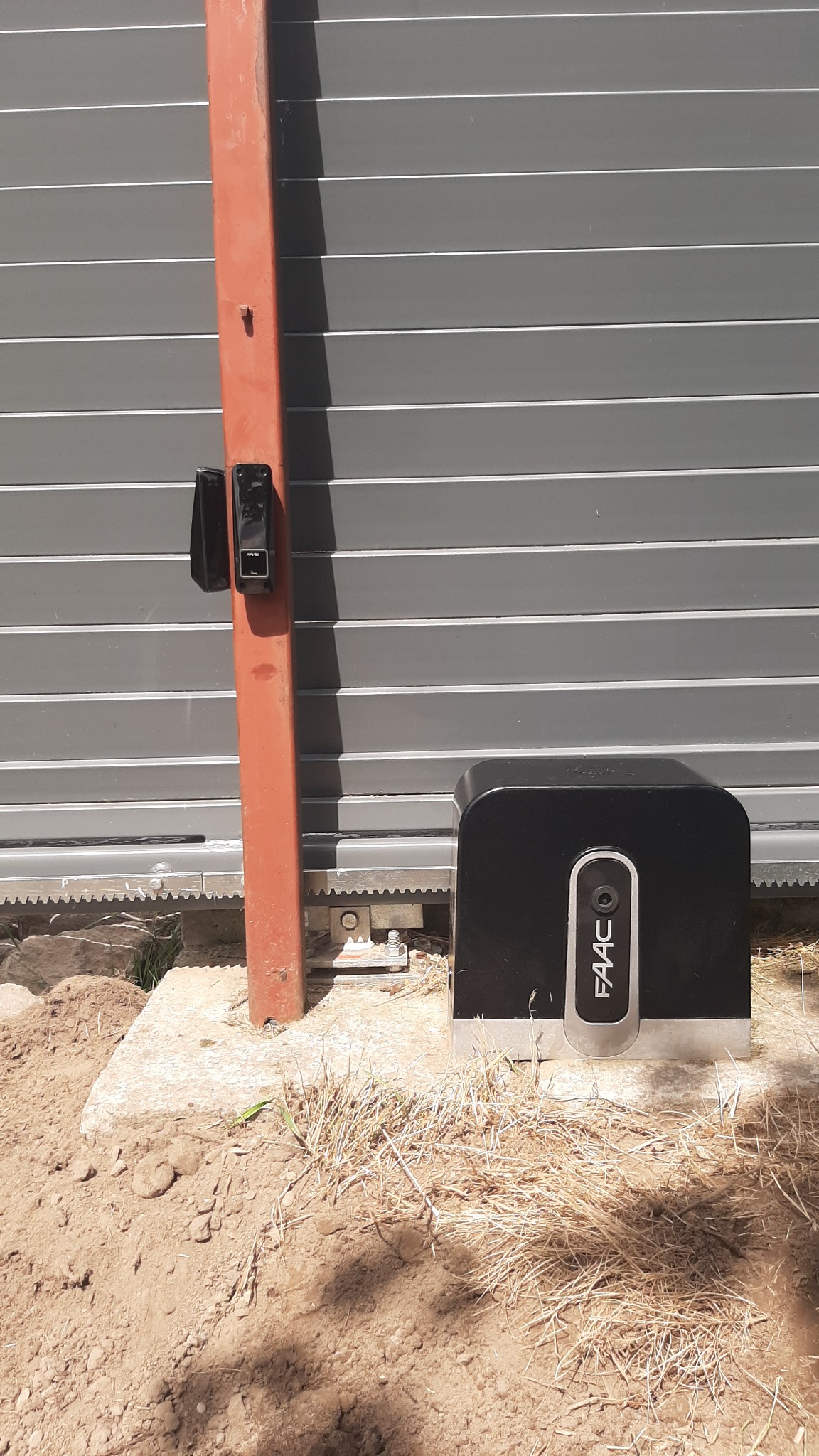 Szara brama przesuwna z pomarańczowym słupkiem i czarnym napędem FAAC na betonowym fundamencie w ziemi.