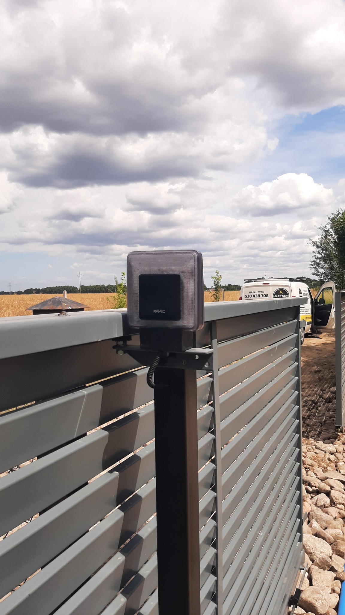Zamontowany napęd FAAC na szarej, metalowej bramie posesji z widocznym w tle polem i firmowym samochodem serwisowym.