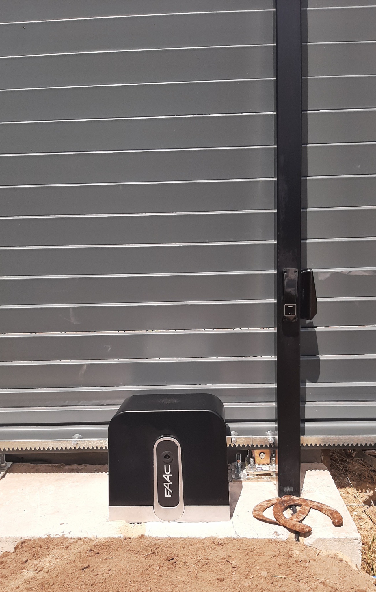 Szara brama przesuwna z zamontowanym czarnym napędem FAAC na betonowej podstawie, obok leży zardzewiały element metalowy.