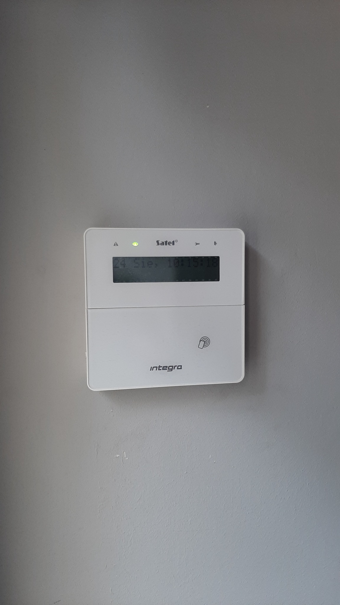 Biała centrala alarmowa Satel Integra zamontowana na szarej ścianie, widoczny wyświetlacz z datą i godziną oraz logo producenta.