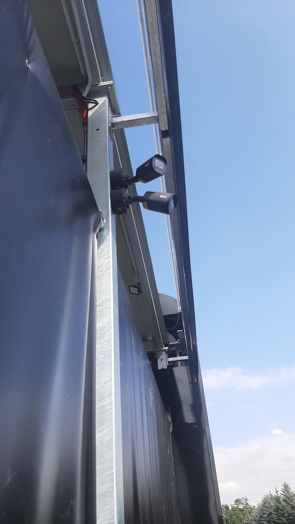 Dwa czarne reflektory przemysłowe zamontowane na metalowej konstrukcji bramy garażowej pod kątem w górę, widoczne przewody i elementy sterujące, błękitne niebo w tle.