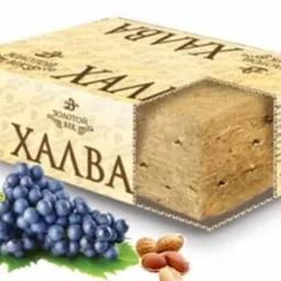 Halwa w kartonowym opakowaniu z wyciętym fragmentem ukazującym strukturę, obok kiść ciemnych winogron i orzeszki.