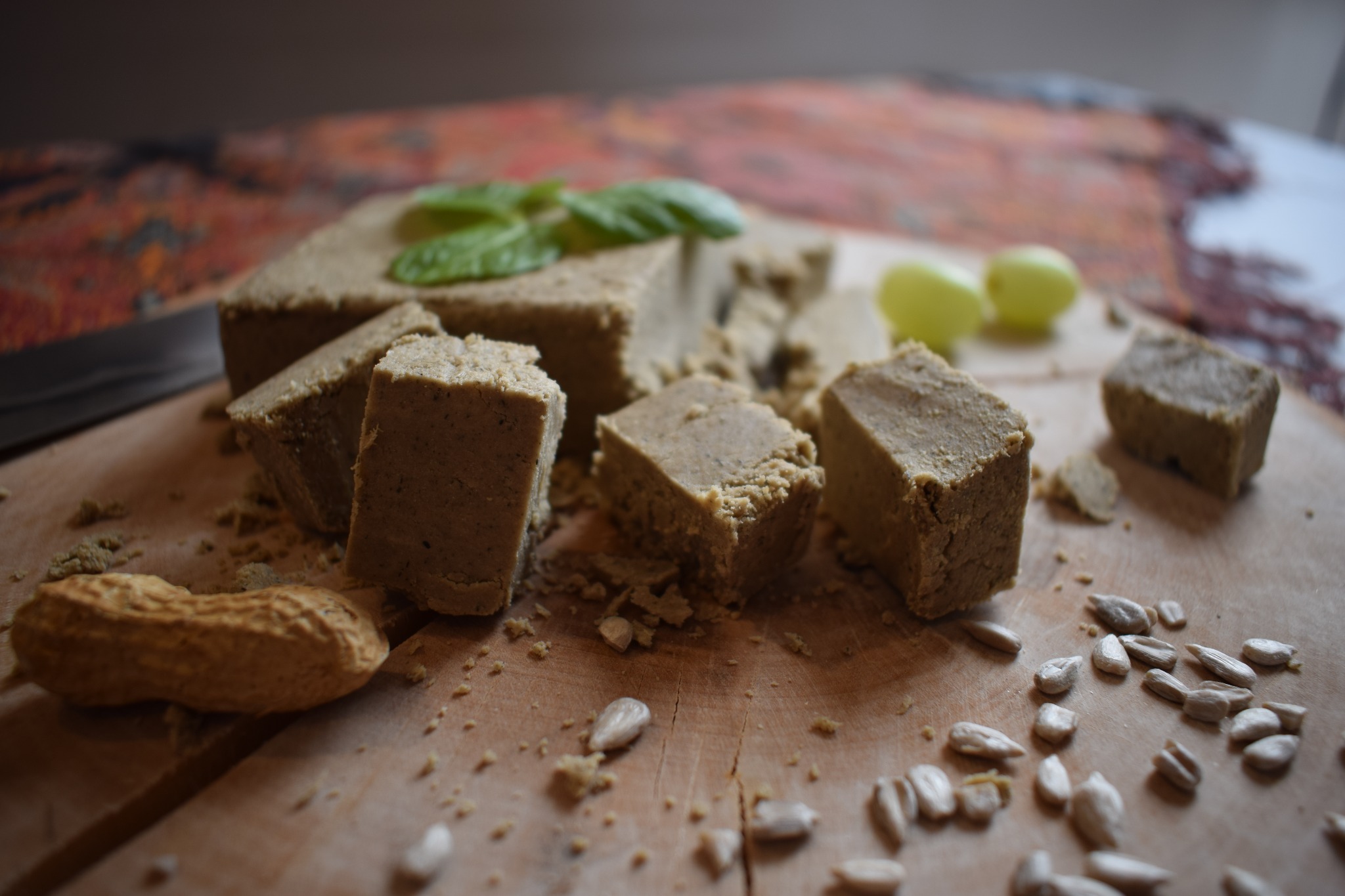 Chałwa słonecznikowa pokrojona w kostki, udekorowana listkami bazylii i winogronami, otoczona orzeszkami ziemnymi i nasionami słonecznika na drewnianej desce.