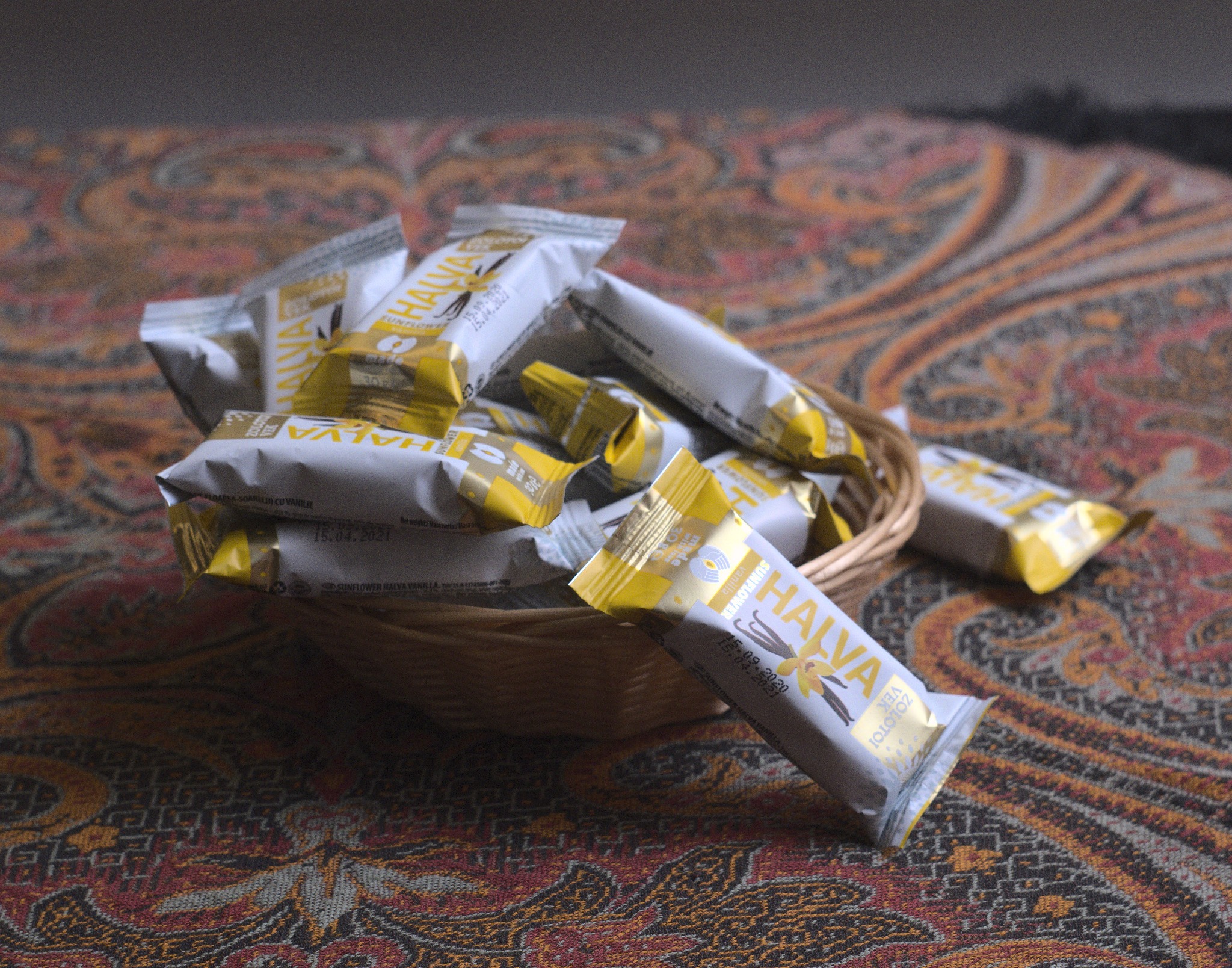 Kosz wiklinowy wypełniony porcjowanymi chałwami słonecznikowymi w opakowaniach z motywem wanilii, leżący na wzorzystej tkaninie.