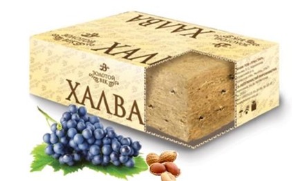 Halwa w kartonowym opakowaniu z wyciętym fragmentem ukazującym strukturę, obok kiść ciemnych winogron i orzeszki.