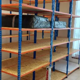 Metalowe regały magazynowe z niebieskimi słupkami i pomarańczowymi poprzeczkami, wypełnione płytami OSB, częściowo załadowane zapakowanymi towarami.