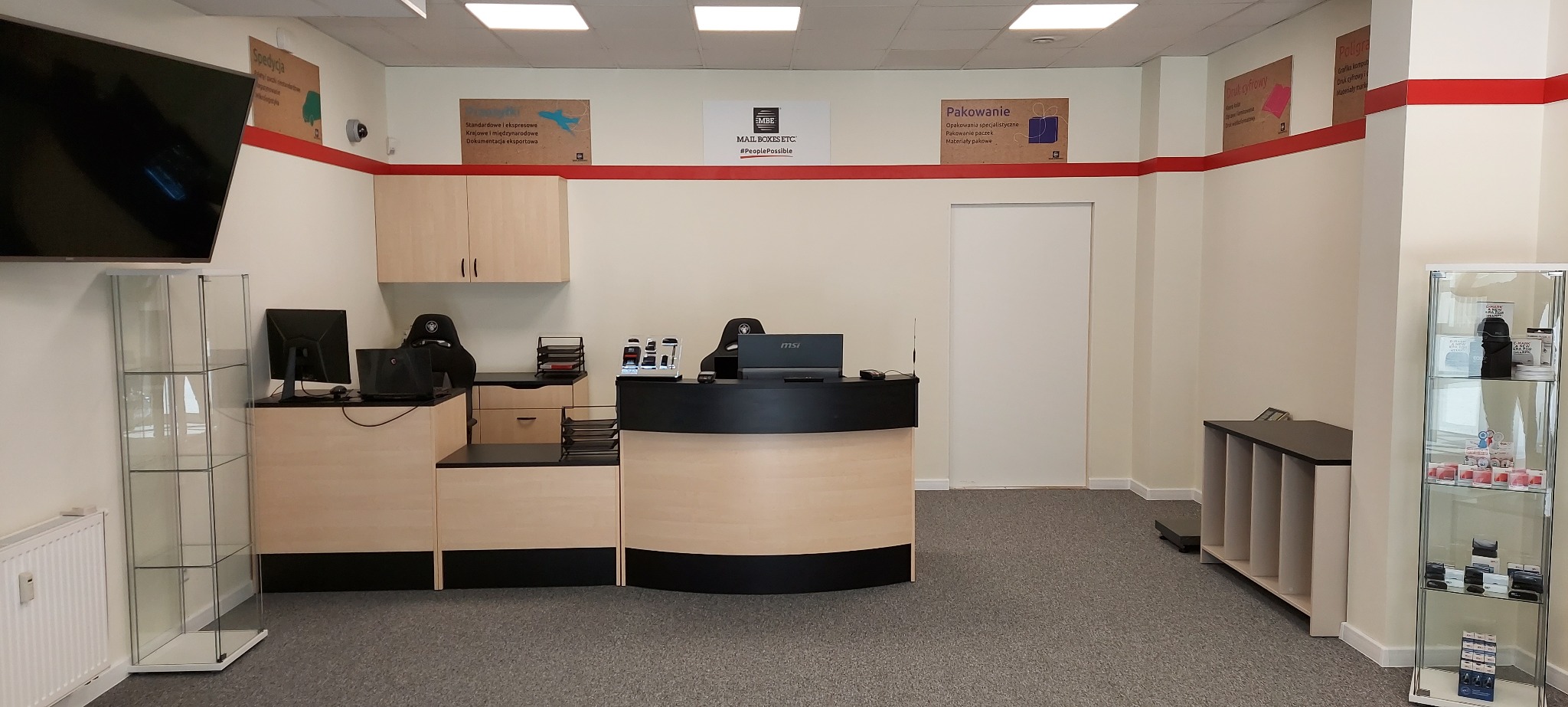 Wnętrze punktu obsługi klienta firmy kurierskiej z ladą recepcyjną, komputerami, regałami i plakatami informacyjnymi o usługach spedycyjnych i materiałach pakowych.