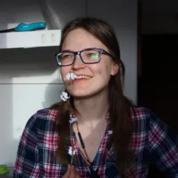 Uśmiechnięta kobieta w okularach i flanelowej koszuli trzymająca w dłoni gałązkę z papierowymi kwiatami, w tle fragment kuchni.