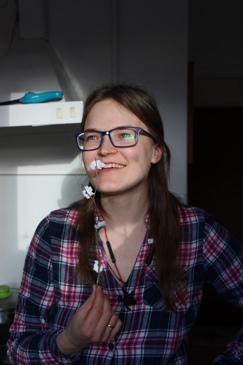 Uśmiechnięta kobieta w okularach i flanelowej koszuli trzymająca w dłoni gałązkę z papierowymi kwiatami, w tle fragment kuchni.