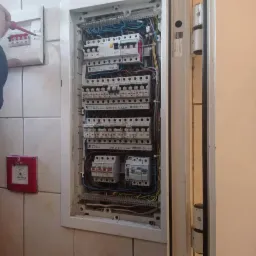 Otwarta skrzynka rozdzielcza z bezpiecznikami i przewodami, widoczny fragment dłoni z czerwonym śrubokrętem, obok czujnik pożarowy na kafelkowanej ścianie.