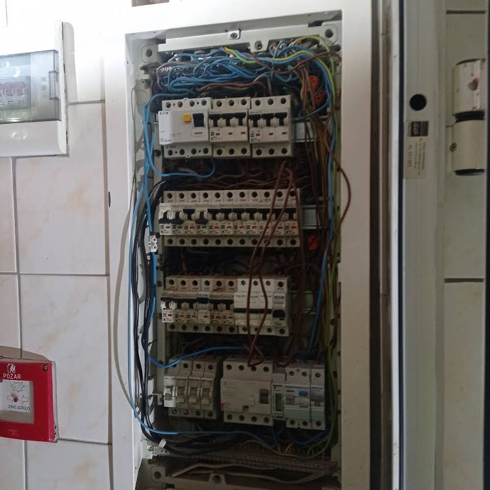 Otwarta skrzynka elektryczna z widocznymi bezpiecznikami, przewodami w kolorach niebieskim, brązowym i zielonym, zamontowana na kafelkowanej ścianie, z czerwonym przyciskiem alarmowym POŻAR po...