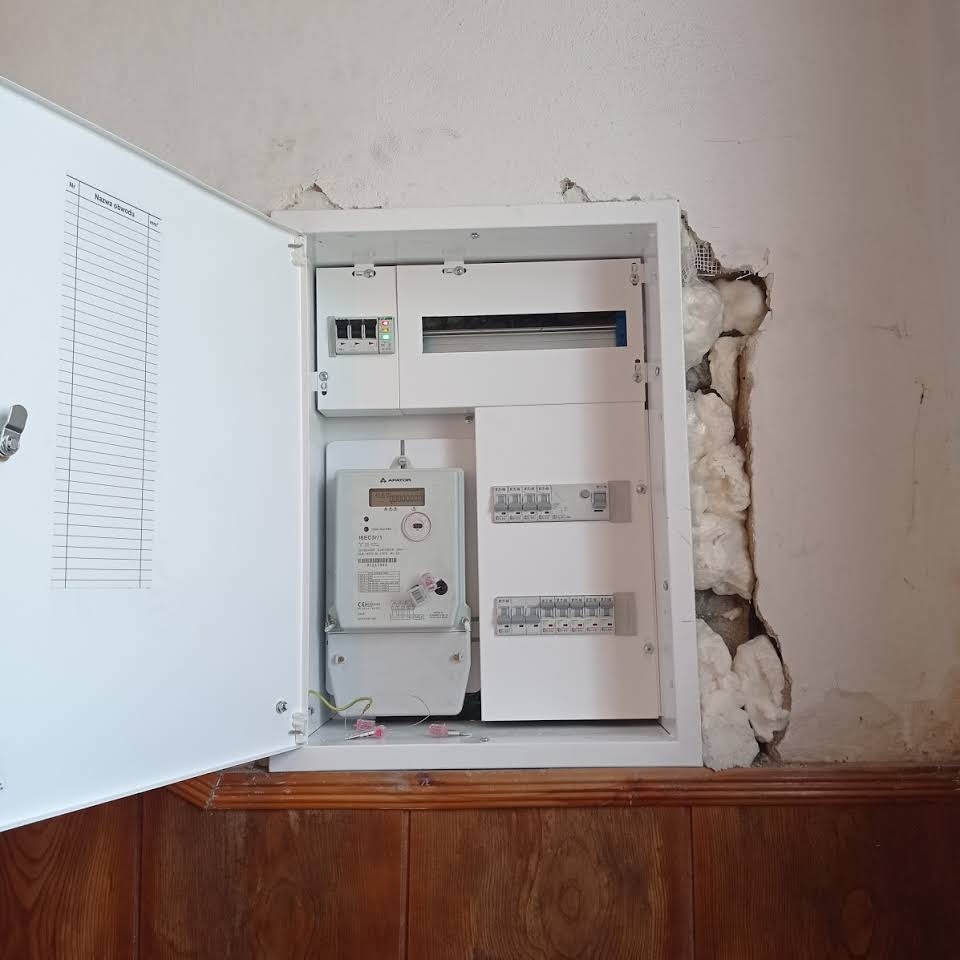 Otwarta skrzynka z bezpiecznikami i licznikiem energii elektrycznej, widoczne zabezpieczenia i instalacja w ścianie z pianki montażowej, drewniana boazeria poniżej.