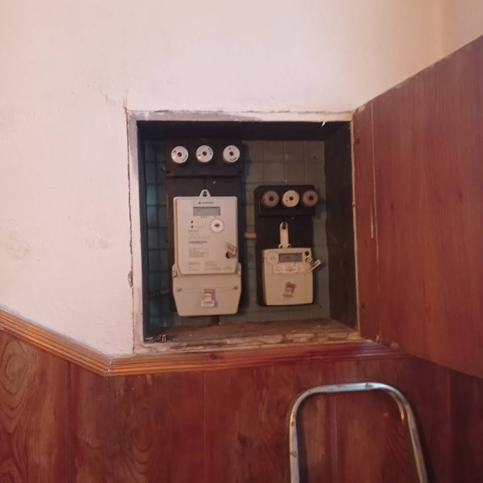 Dwa liczniki energii elektrycznej zamontowane w skrzynce w ścianie, widoczne bezpieczniki topikowe i fragment drabiny w dolnej części kadru.