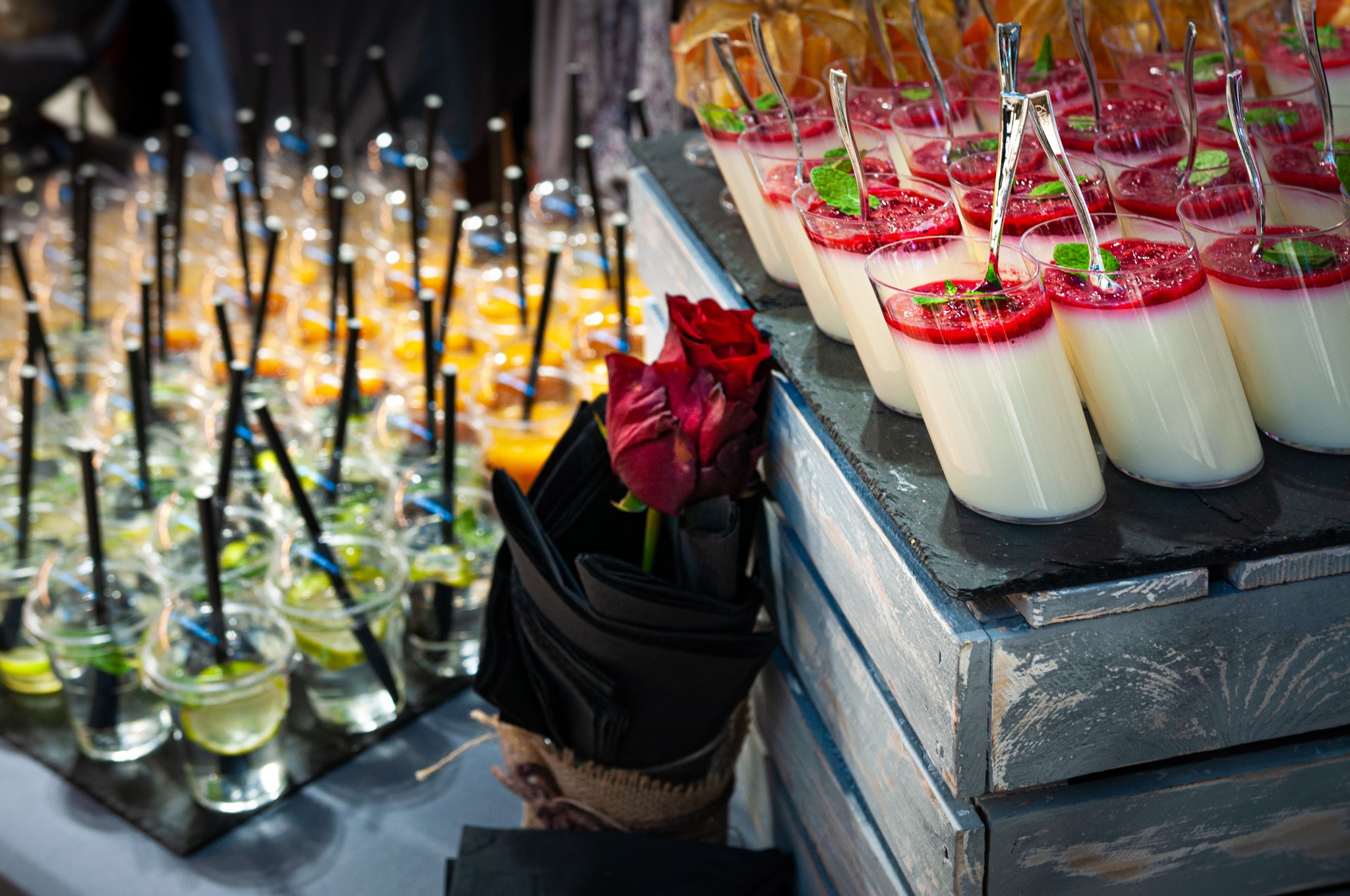 Elegancko przygotowany bufet z deserami w szklanych naczyniach, udekorowanymi czerwoną galaretką, miętą i srebrnymi łyżeczkami; obok drinki z limonką i słomkami, a w centrum bukiet ciemnoczerwonych...