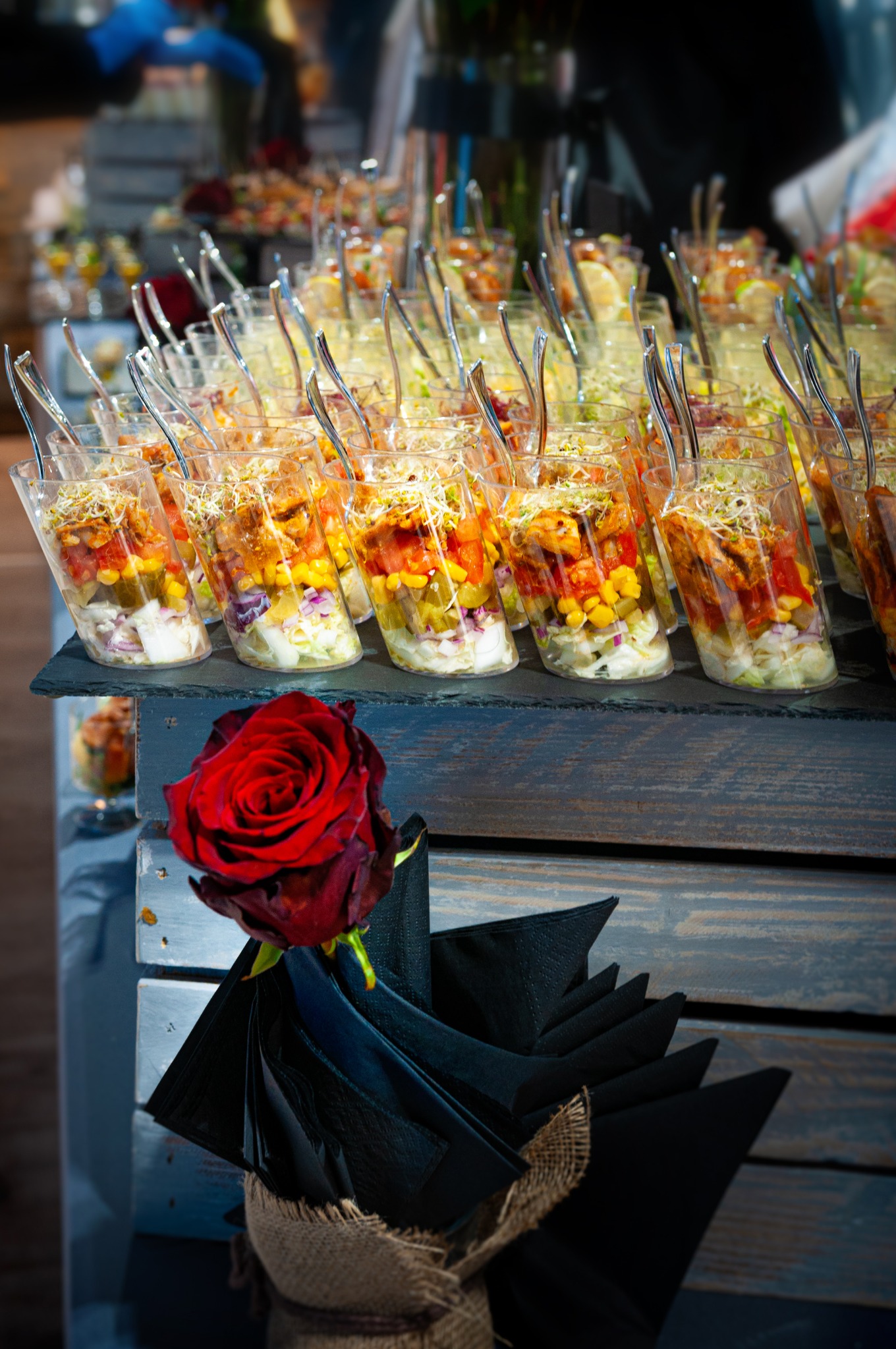 Szereg przezroczystych kubeczków z sałatką warstwową, zawierającą sałatę, kukurydzę, pomidory i kiełki, ustawionych na ciemnej, drewnianej powierzchni, z pojedynczą czerwoną różą i czarnymi...