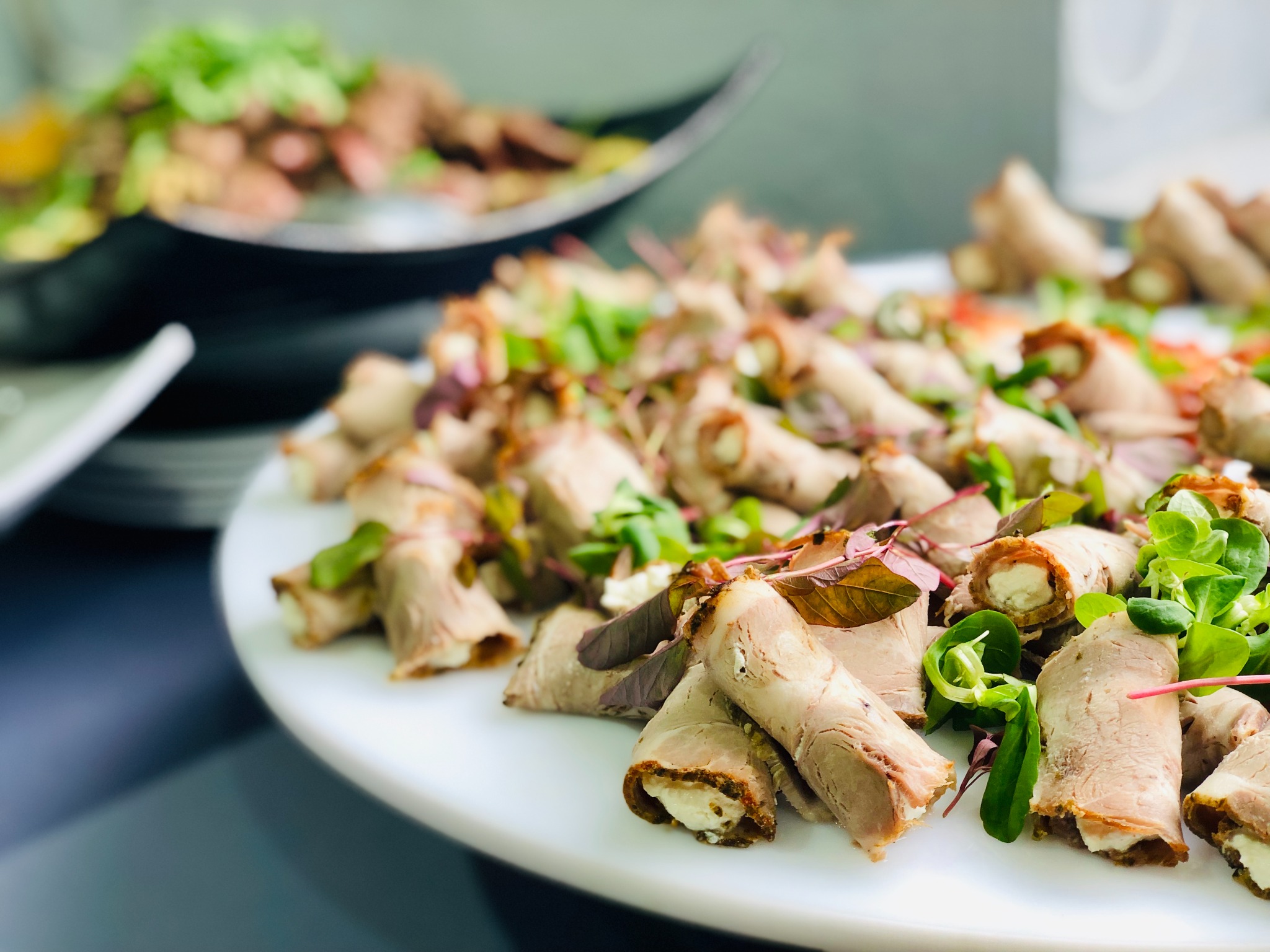 Elegancko ułożone roladki z mięsa z kremowym nadzieniem, udekorowane świeżymi listkami sałat na białym półmisku, w tle widoczna kolejna potrawa z zielenią.