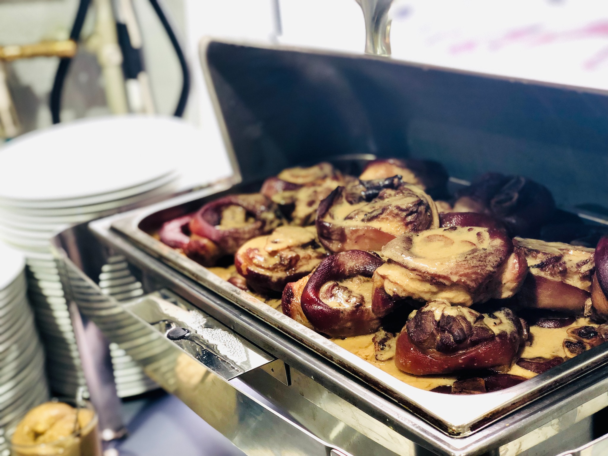 Szynki wieprzowe z sosem w metalowym podgrzewaczu cateringowym, obok stos białych talerzy.