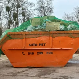 Pomarańczowy kontener z czarnym napisem AUTO-MET i numerem telefonu, wypełniony gruzem i zabezpieczony zieloną siatką, na tle drzew bez liści i pochmurnego nieba.