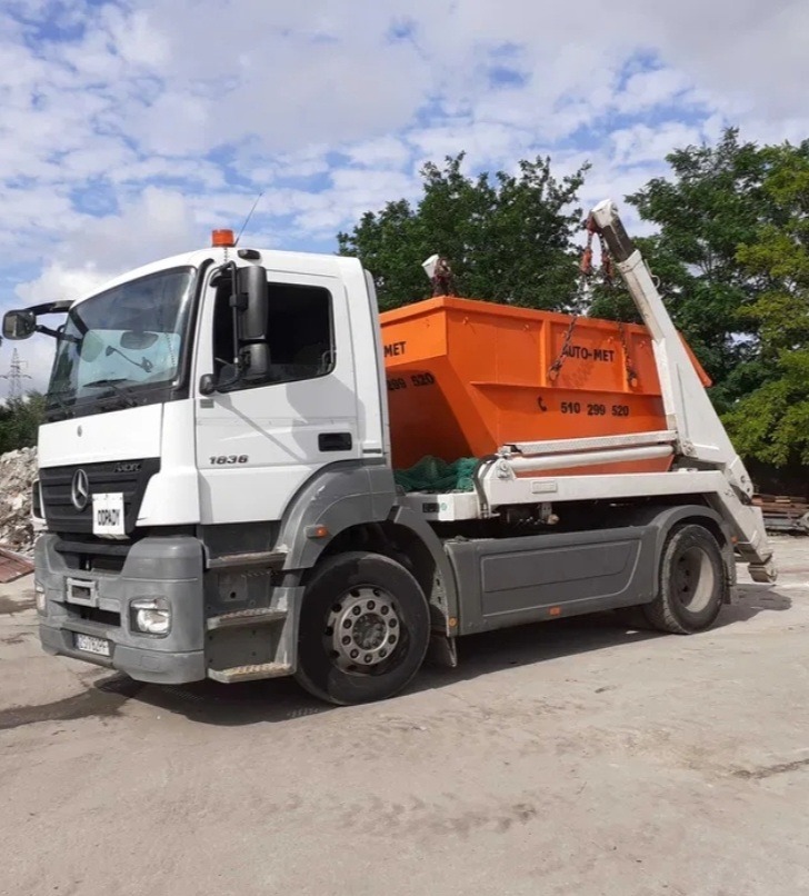 Biała ciężarówka Mercedes z pomarańczowym kontenerem na gruz Auto-Met na betonowym placu, widoczne drzewa w tle, dzień z lekkim zachmurzeniem.