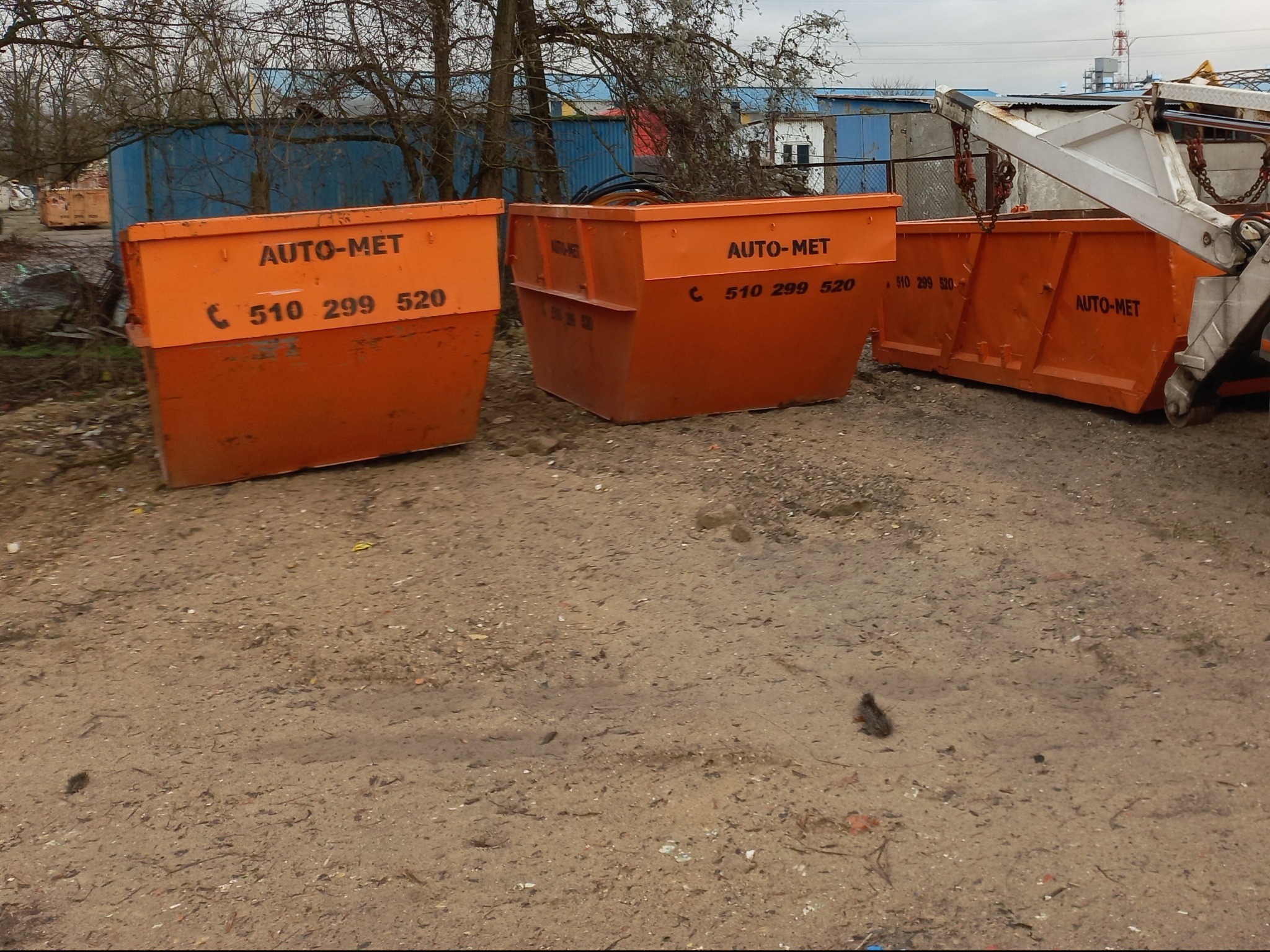 Pomarańczowe kontenery z napisem AUTO-MET i numerem telefonu stoją na ziemi na placu budowy z widocznymi budynkami gospodarczymi w tle.