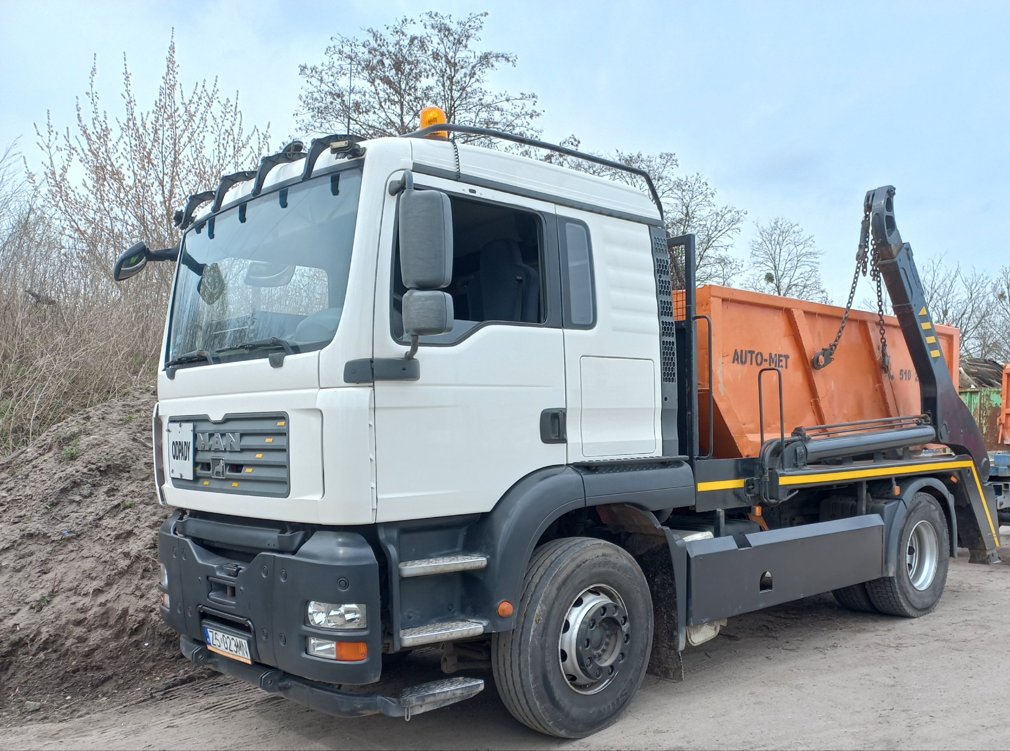 Biała ciężarówka MAN z pomarańczowym kontenerem na gruz, zaparkowana na szutrowej drodze, z widocznym stosem ziemi w tle i drzewami bez liści.
