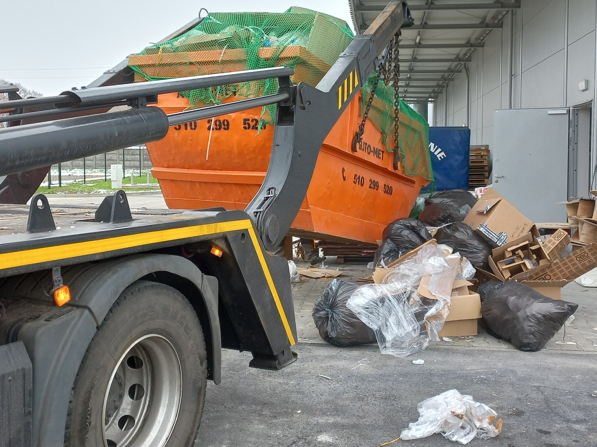 Pomarańczowy kontener na gruz z zieloną siatką, podnoszony przez ramię samochodu ciężarowego, obok porozrzucane kartony i worki na śmieci na betonowym placu.