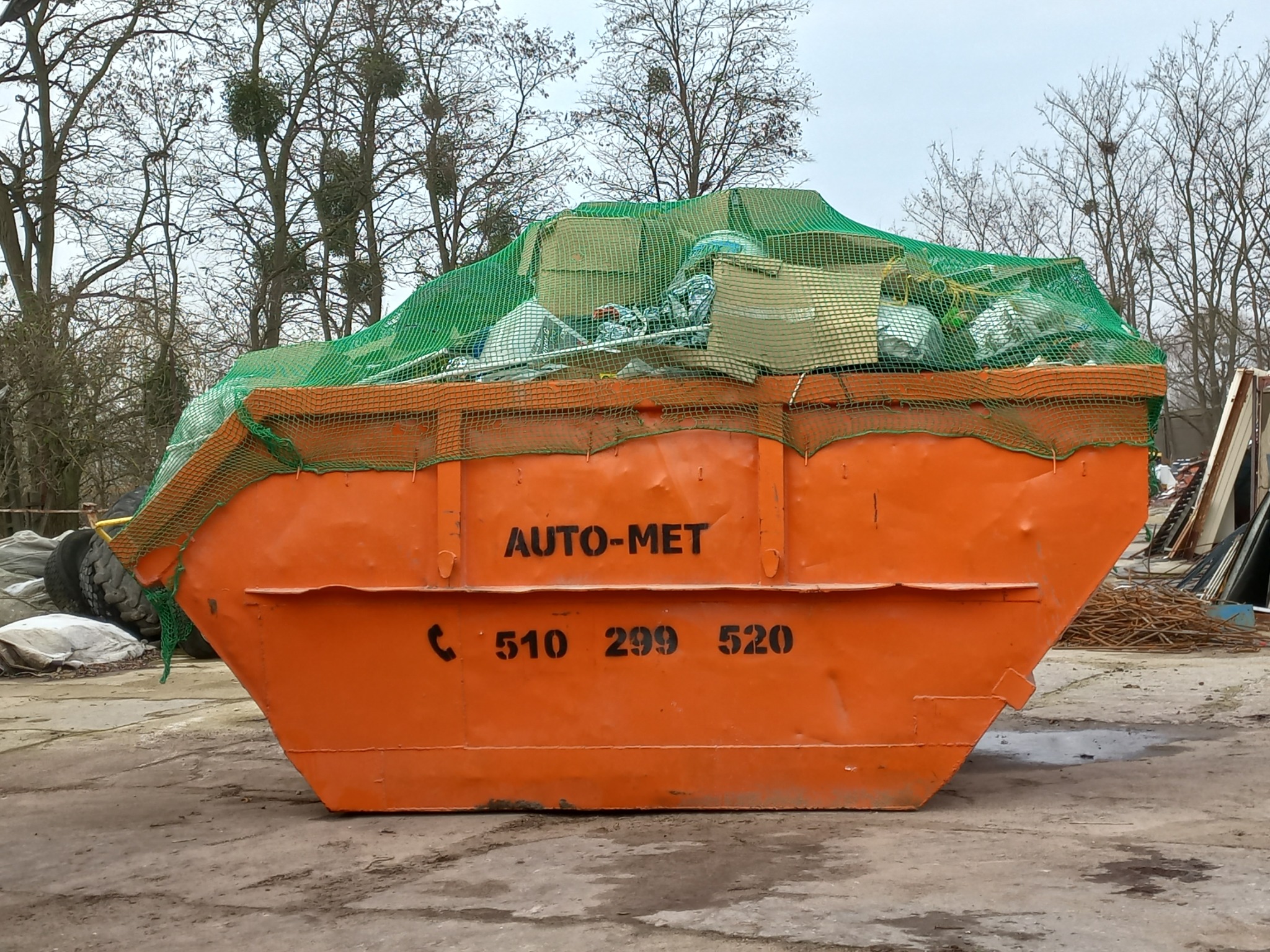Pomarańczowy kontener z czarnym napisem AUTO-MET i numerem telefonu, wypełniony gruzem i zabezpieczony zieloną siatką, na tle drzew bez liści i pochmurnego nieba.