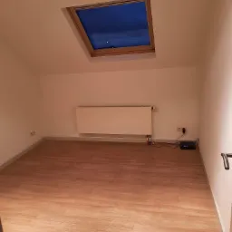 Poddasze z oknem dachowym, panelami podłogowymi imitującymi drewno i białym grzejnikiem na ścianie.