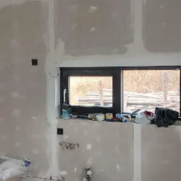 Wnętrze w trakcie prac wykończeniowych: ściany pokryte gładzią gipsową, widoczne puszki elektryczne, okno z widokiem na składowane drewno na zewnątrz, narzędzia i materiały budowlane na parapecie...